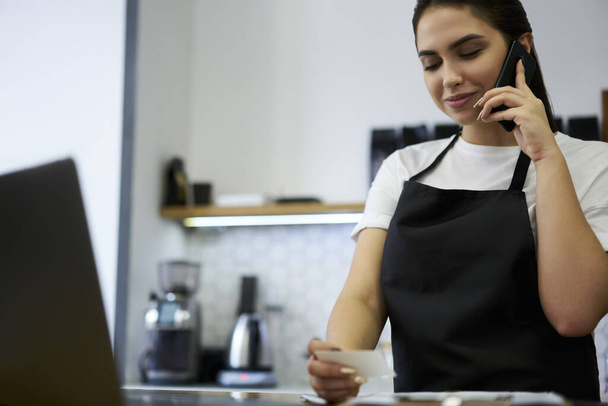 Professional manager of restaurant enjoying job making mobile phone call to delivery service, positive 20s woman barista making booking via cellular holding visit card standing at working space - Фото, зображення