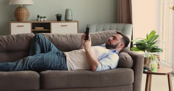 Relaxed millennial guy lie on couch watch video on phone - Záběry, video