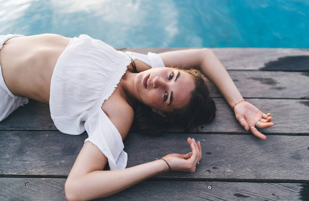 High angle of sensual female in white top lying on poolside and looking at camera while enjoying vacation in summer - Фото, изображение