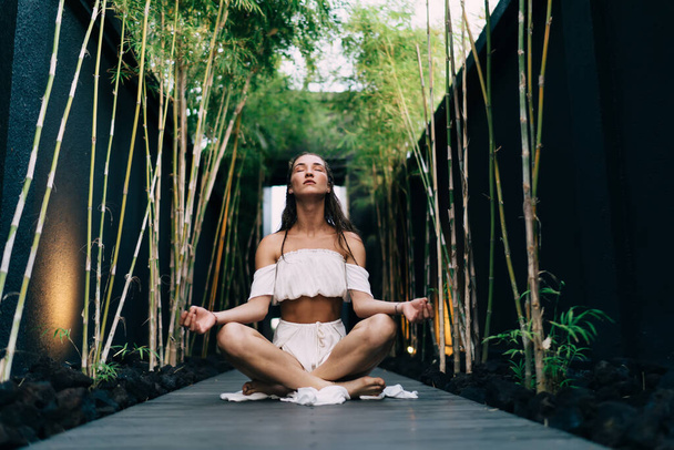 Low angle of tranquil female in trendy clothes sitting on wooden pathway in Padmasana position during yoga session in open air - Zdjęcie, obraz