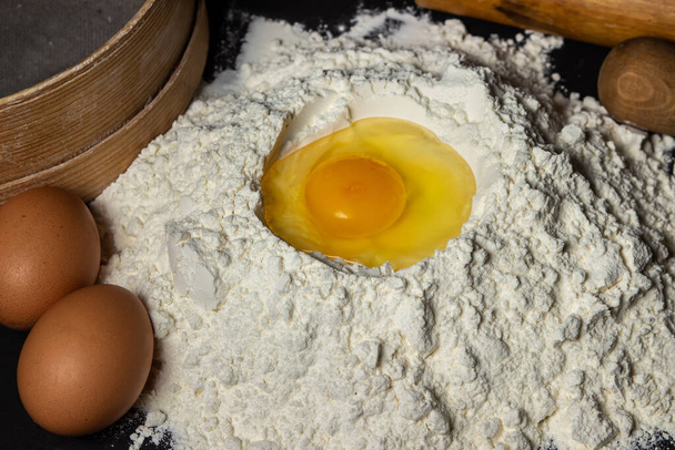 Raw egg in flour on a black background. Dough preparation. homemade baking - 写真・画像