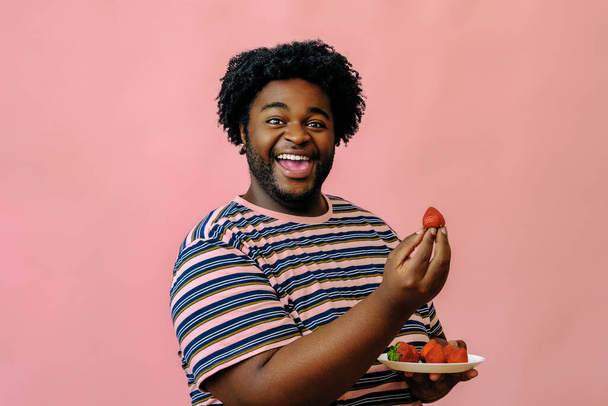 joven feliz afroamericano hombre comiendo fresas en el estudio sobre fondo rosa   - Foto, imagen