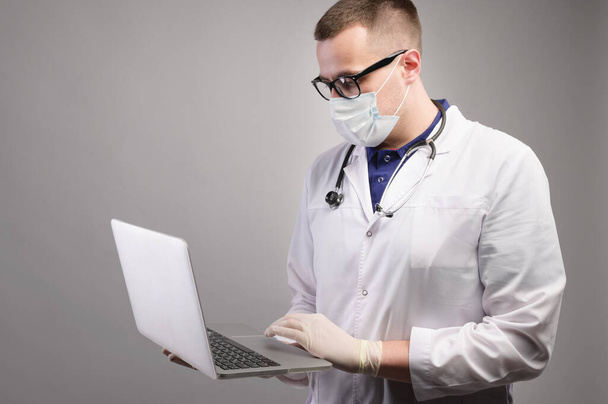 Successful young caucasian male doctor in a white medical uniform and with a stethoscope, busy working on a laptop, typing on a computer, consulting a patient online. Studio portrait - Foto, Bild