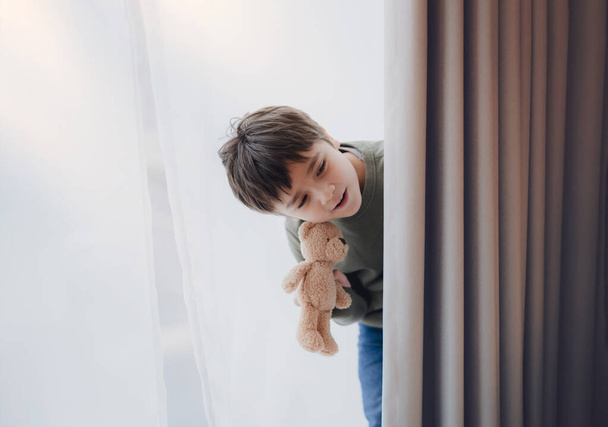 Niño lindo sosteniendo oso de peluche de pie detrás de la cortina de encaje con luz brillante en la mañana, niño feliz con la cara sonriente en el alféizar de la ventana y juega en el escondite detrás de la cortina - Foto, imagen