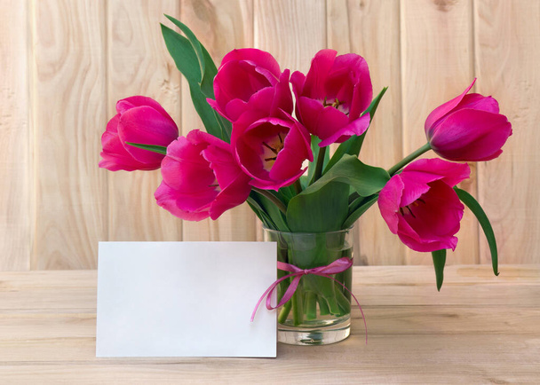 Bouquet pink tulips flowers in glass white vase with white paper card note with space for text on table on a wooden background - Valokuva, kuva