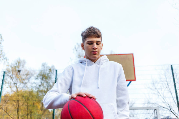 Cute teenager in white hoodie playing basketball. Young boy with red ball learning dribble and shooting on the city court. Hobby for kids, active lifestyle - Foto, Imagem