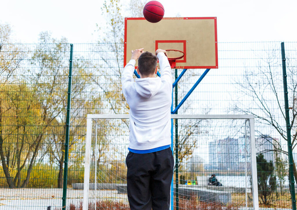Cute teenager in white hoodie playing basketball. Young boy with red ball learning dribble and shooting on the city court. Hobby for kids, active lifestyle - Foto, Imagen