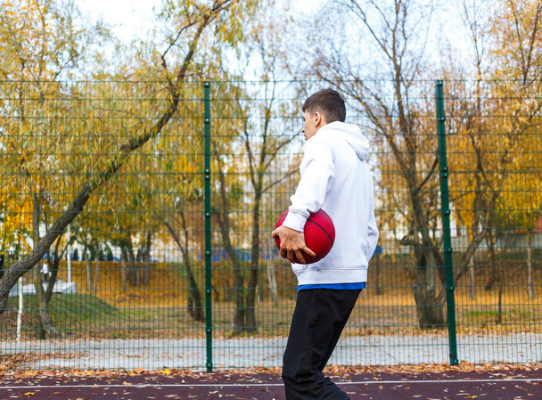 Cute teenager in white hoodie playing basketball. Young boy with red ball learning dribble and shooting on the city court. Hobby for kids, active lifestyle - Foto, afbeelding