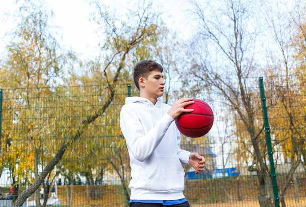 Cute teenager in white hoodie playing basketball. Young boy with red ball learning dribble and shooting on the city court. Hobby for kids, active lifestyle - Фото, зображення