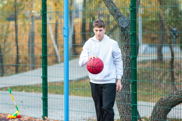 Cute teenager in white hoodie playing basketball. Young boy with red ball learning dribble and shooting on the city court. Hobby for kids, active lifestyle - Valokuva, kuva