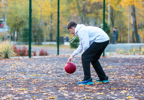 Cute teenager in white hoodie playing basketball. Young boy with red ball learning dribble and shooting on the city court. Hobby for kids, active lifestyle - Foto, Imagem