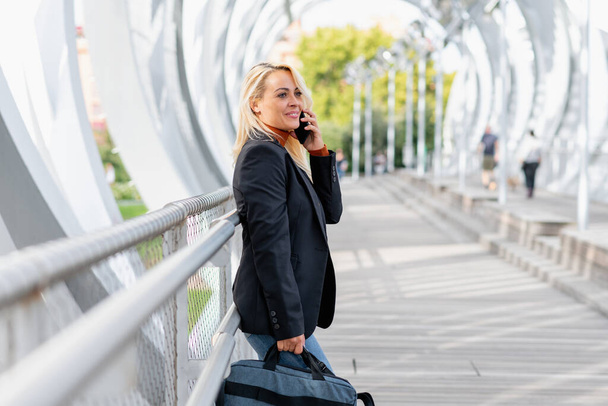 Smiling woman leaning on a railing of a urban bridge while talking with the mobile carrying a bag - Foto, imagen