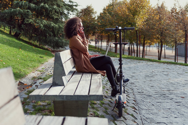 mujer sentada en un banco del parque habla por teléfono tiene un ordenador portátil y un scooter eléctrico - Foto, Imagen