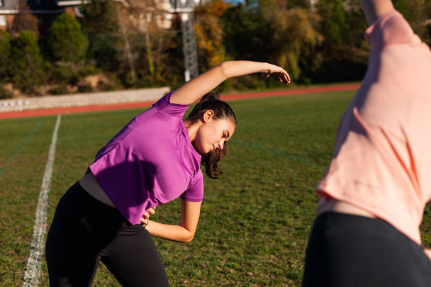 dos mujeres jóvenes haciendo estiramiento en la hierba de una pista de atletismo antes de practicar deportes - Foto, Imagen