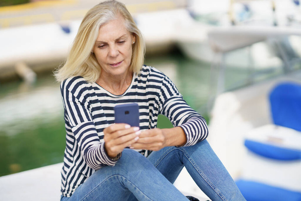 海の港に座っているスマートフォンを使っているシニア女性. - 写真・画像