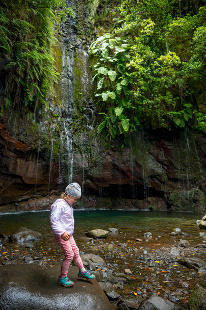 Traveler girl wearing sport jacket walking on shores of waterfall. Travel lifestyle. View from back. - Foto, afbeelding