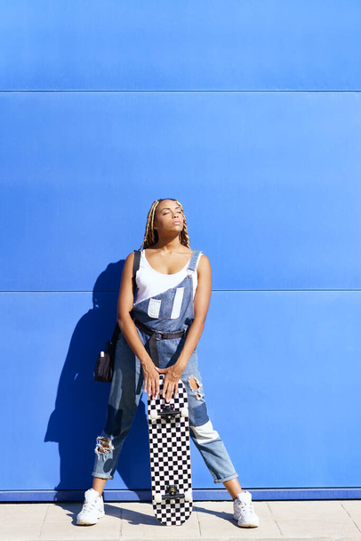黒の女の子は、青い壁の背景にスケートボードで、カジュアルな服装. - 写真・画像