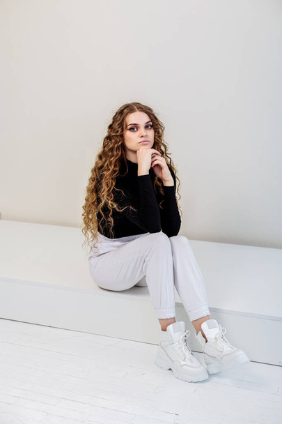 A beautiful woman with long curly hair in a black sweater, white pants, white leather sneakers. New collection of women's casual shoes - 写真・画像