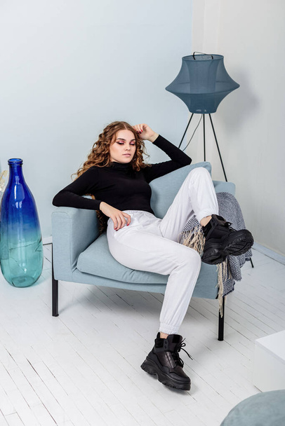 A beautiful woman with long curly hair in a black sweater, white pants, black leather sneakers. New collection of women's casual shoes - Valokuva, kuva