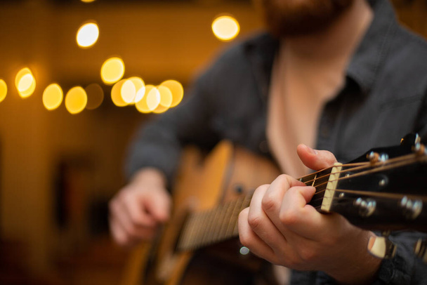 A young guy with a beard plays an acoustic guitar in a room with warm lighting - Foto, imagen
