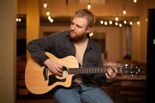 Un joven con barba toca una guitarra acústica en una habitación con una cálida iluminación - Foto, Imagen