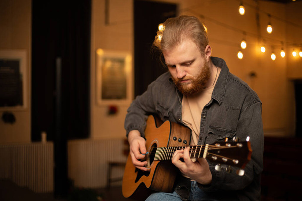 A young guy with a beard plays an acoustic guitar in a room with warm lighting - 写真・画像