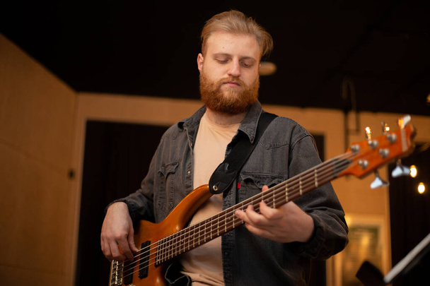 A young guy with a beard plays a bass guitar with five strings - Zdjęcie, obraz