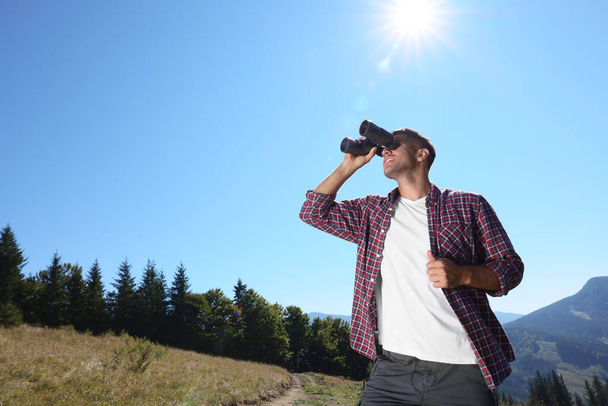 Man looking through binoculars in mountains on sunny day, low angle view - Foto, imagen