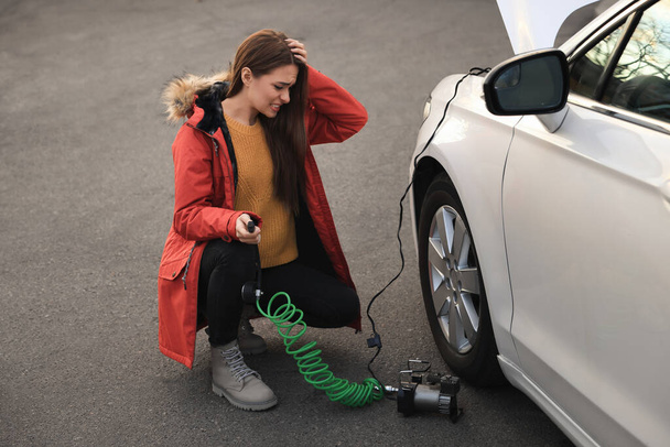 Young woman trying to inflate car tire with air compressor on street - Foto, afbeelding