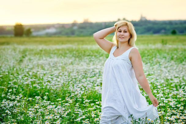 Eine junge mollige schöne Frau ruht sich bei Sonnenuntergang auf einem Kamillenfeld aus. Eine Plus-Size-Frau im weißen Kleid auf einer Wiese mit Gänseblümchen . - Foto, Bild