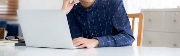 Młody Azjata rozmawiający przez telefon i pracujący w domu z laptopem, wolny strzelec korzystający z notebooka do Internetu online i mówiący na smartfonie, w życiu domowym, w biznesie i komunikacji. - Zdjęcie, obraz