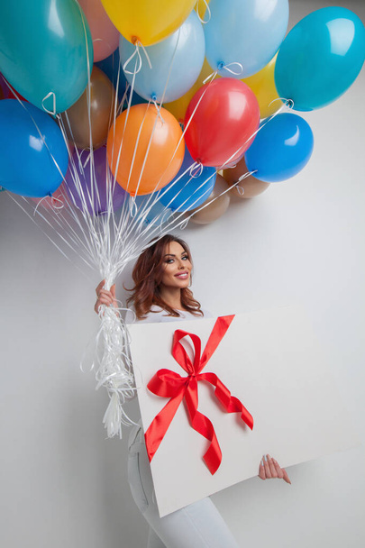 Young smiling woman with many colorful balloons and big gift box - 写真・画像