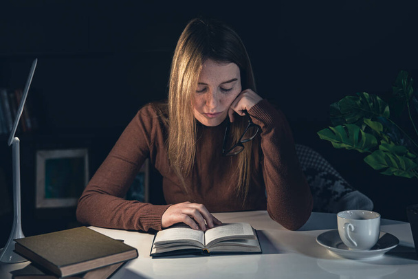 Молодая женщина сидит за столом и читает ночью книгу с лампой. - Фото, изображение
