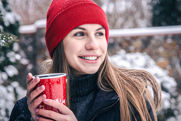 Une jeune femme boit une boisson chaude dans une tasse thermique rouge en hiver. - Photo, image
