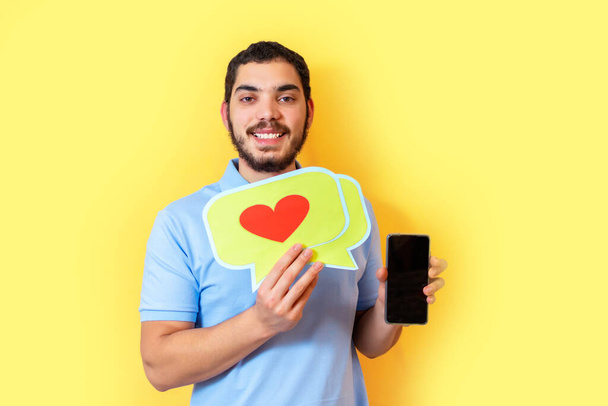 Young man holds up a speech balloon with a heart and shows the smart phone blank screen. Social media concept. - Fotó, kép