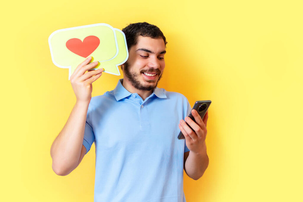 Young man holds up a speech balloon with a heart and used the smart phone. Social media concept. - Фото, изображение