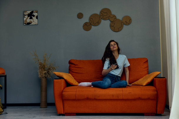 mujer sonriente charlando en el sofá naranja con un teléfono inteligente tecnologías - Foto, Imagen