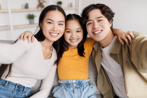 Smiling teenager korean girl, millennial woman and man make family selfie on gadget in minimalist living room - Photo, Image