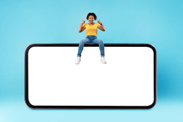 Positive millennial black woman showing thumb up gesture, sitting on top of huge smartphone over blue background, mockup - Фото, изображение