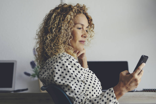 Pretty adult woman use mobile phone connection to call and work sitting at the office - modern business female people with technology device - laptop in background on the desktop - job occupation lifestyle - lady with blonde curly hair - Valokuva, kuva