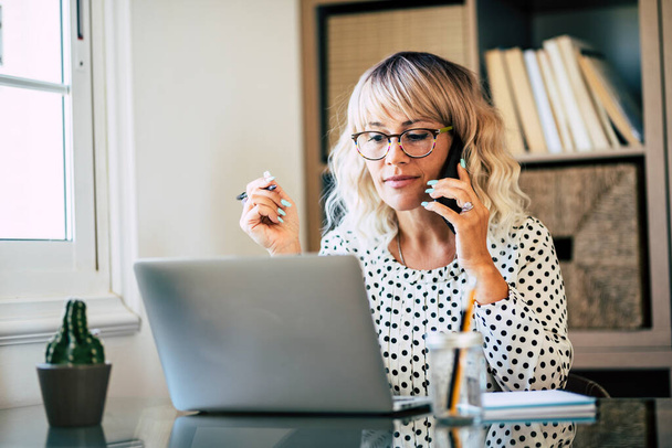 Young adult entreprenaur woman working at office with laptop computer and mobile phone cell connection - professional business lady with eyewear busy in smart working at home - modern people online job - Zdjęcie, obraz