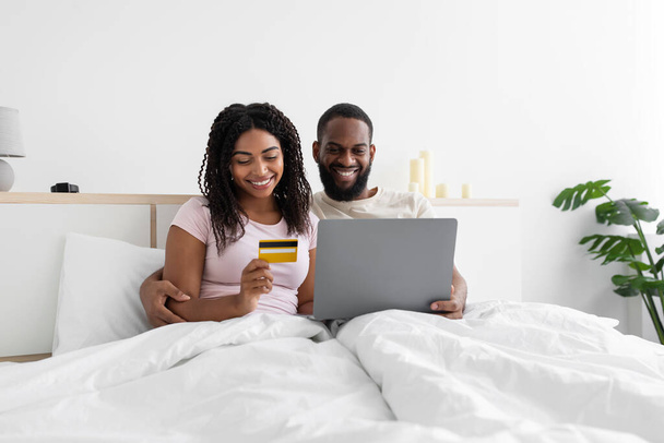 Happy young black woman and husband with computer and credit card sitting on bed in bedroom interior - Fotoğraf, Görsel