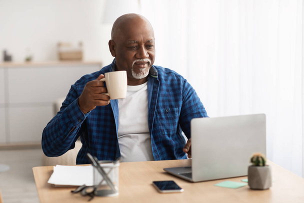 negro maduro hombre trabajando en portátil beber café en lugar de trabajo - Foto, imagen