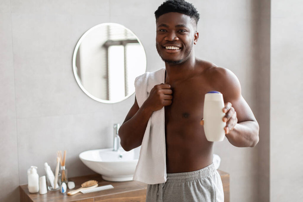 sourire noir mâle montrant douche bouteille de gel debout dans salle de bain - Photo, image