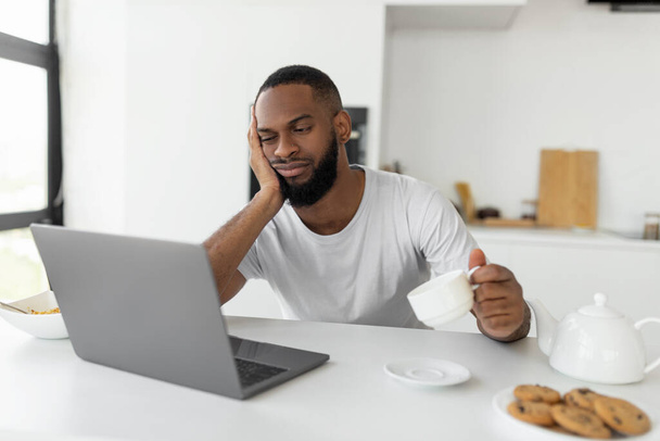 Tired black man using laptop sitting at kitchen table - 写真・画像