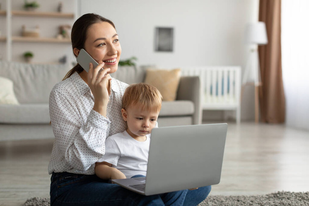 Happy mother with toddler son chatting on smartphone and using laptop computer, sitting on carpet and looking aside - Photo, Image