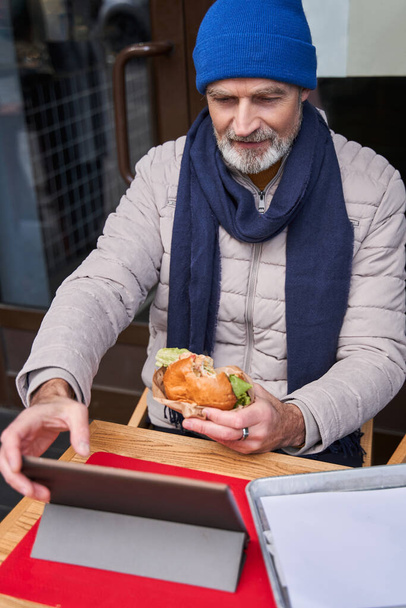 Confident grey haired man eating burger and typing at the tablet - Фото, изображение