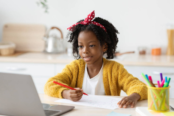 Мила маленька чорна дівчинка малює на кухні під час навчання онлайн з ноутбуком
 - Фото, зображення