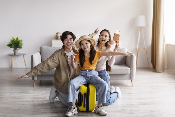 Щаслива дівчина-підліток у капелюсі тримає літак, сидячи на валізі, молода азіатка і чоловік радіють, розважаються і показують паспорт
 - Фото, зображення