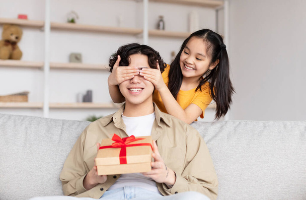 Smiling teen girl closes eyes of young asian man and present box with gift, sit on sofa in living room interior - Foto, Imagen